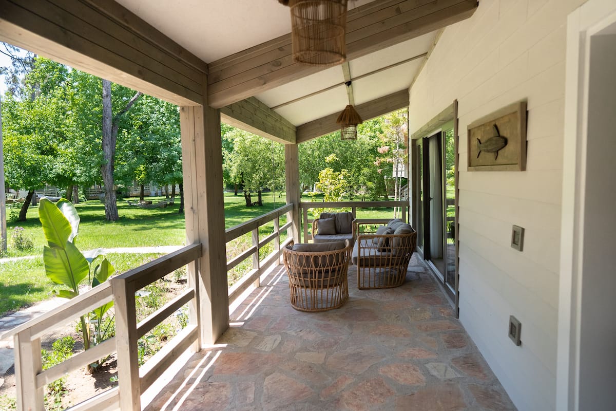 A covered porch with wicker chairs, stone flooring, and a view of a green park.