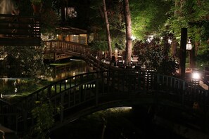 Vista noturna de um jardim japonês tradicional, com uma ponte de madeira sobre um lago, cercado por árvores e iluminado por luzes suaves.
