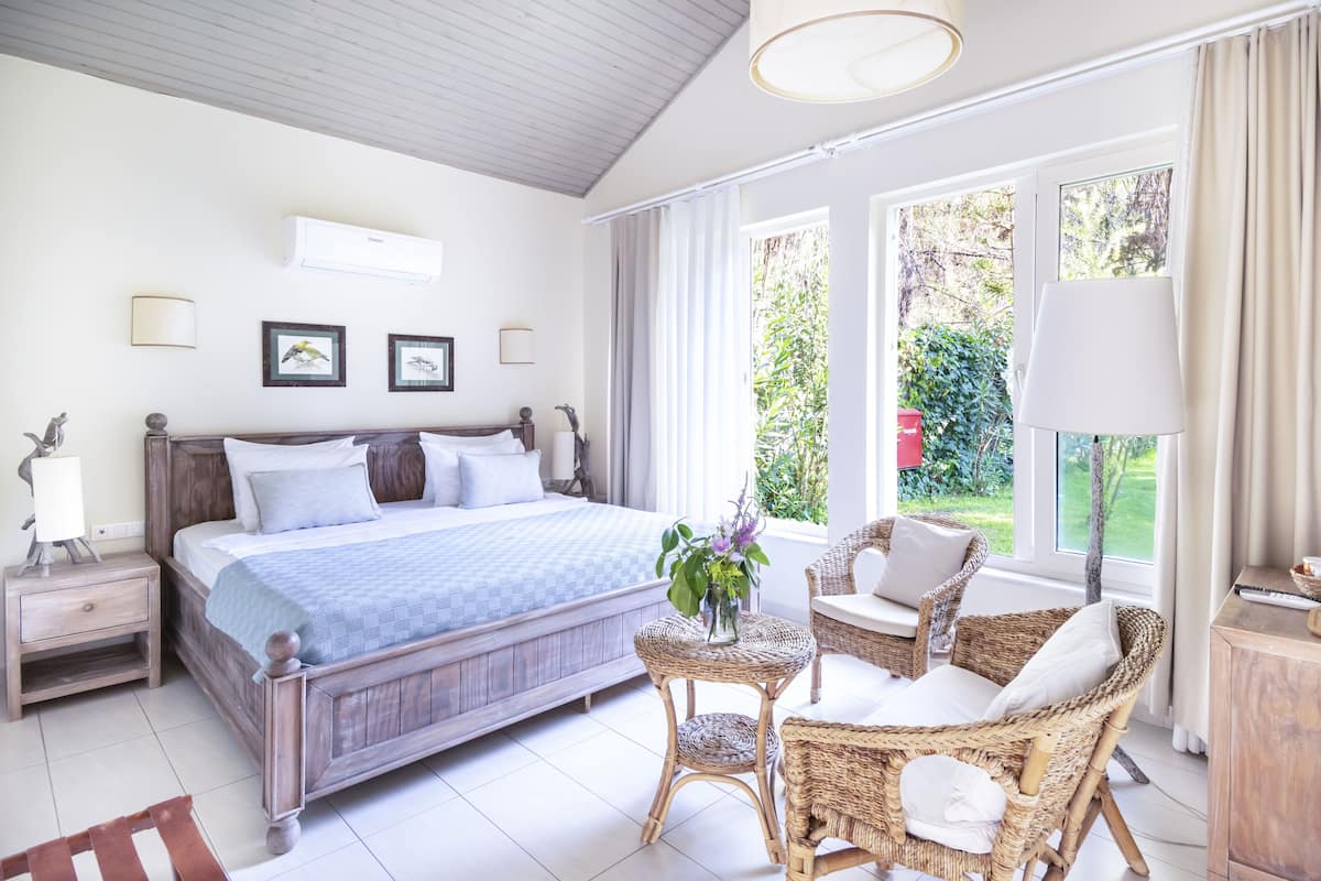 A bedroom with a large bed, a bedside table, a wicker chair, and a window with a view of greenery.