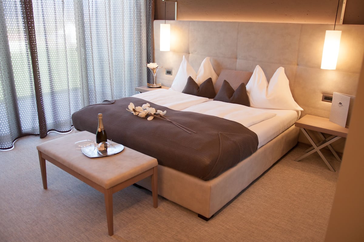 Ein modernes Hotelzimmer mit einem großen Bett, einem Nachttisch mit Flasche und Gläsern sowie einem kleinen Tisch mit einer Vase.