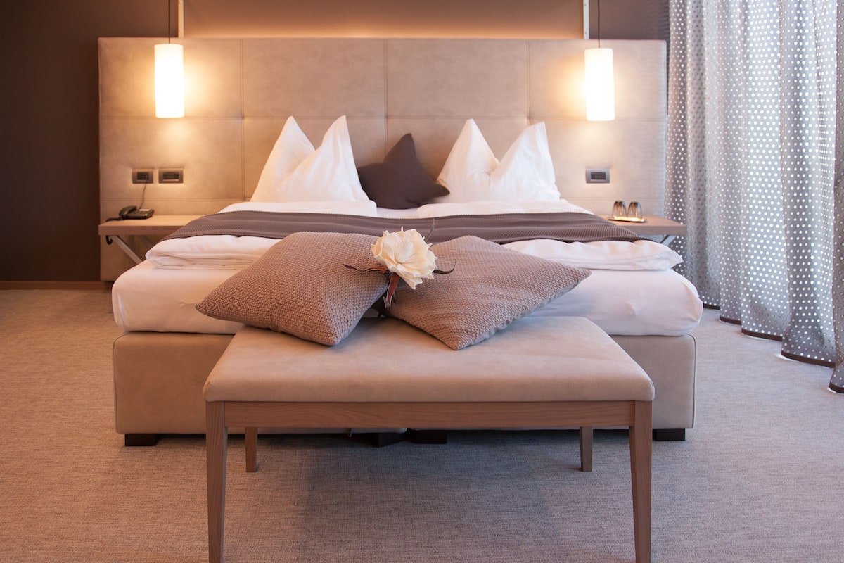 Ein modernes Hotelzimmer mit einem großen Bett, einem Nachttisch mit Lampe und Blick auf die Stadt durch das Fenster.