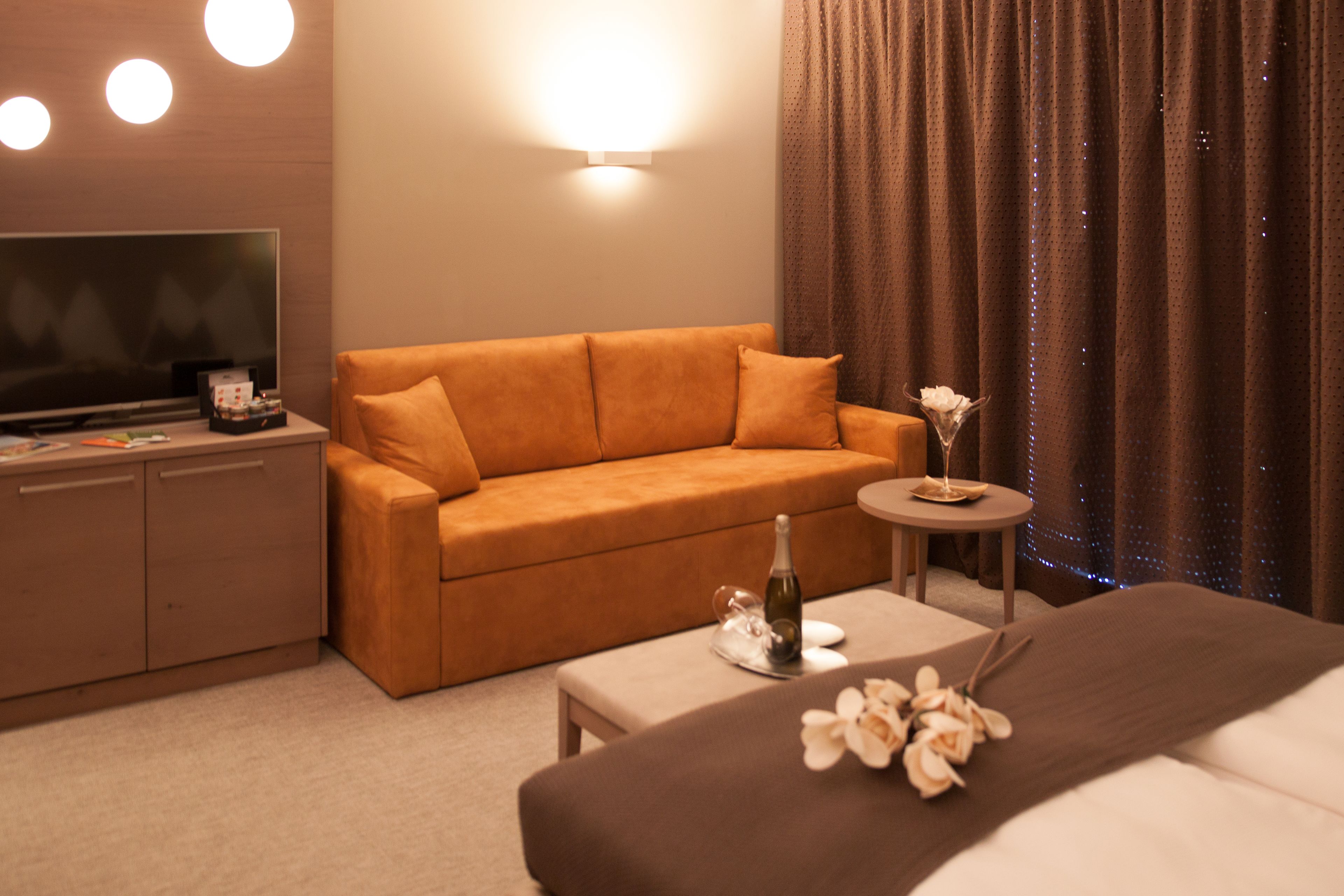 A hotel room with a sofa, a small round table with a bottle and flowers, a TV, and a bed.