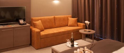 A hotel room with a sofa, a small round table with a bottle and flowers, a TV, and a bed.