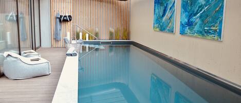 An indoor swimming pool with a wooden deck, a glass wall, and abstract paintings on the wall.