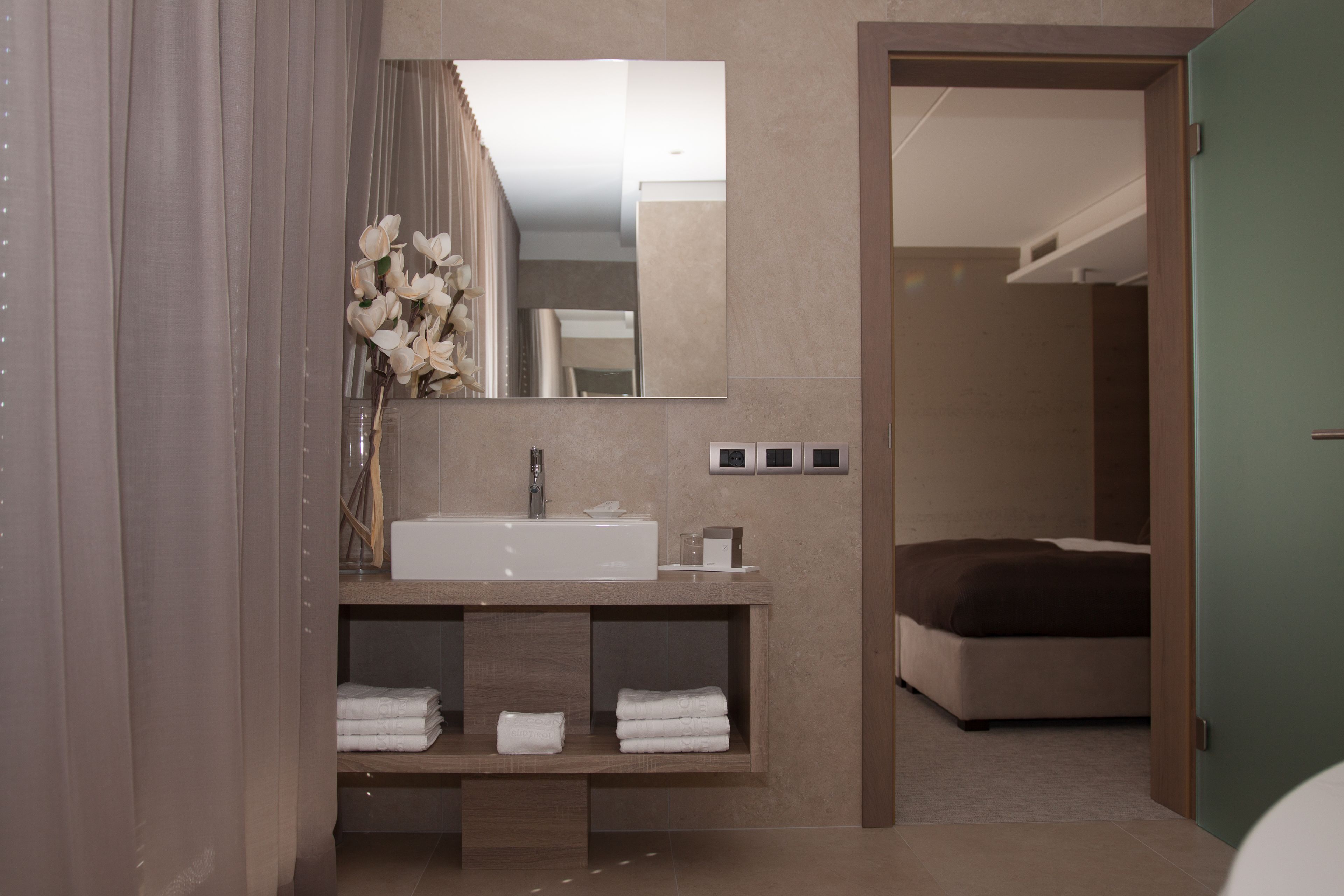 Une salle de bain moderne avec un grand miroir, un lavabo et un meuble-vasque doté d’étagères. On aperçoit un lit dans la pièce adjacente.