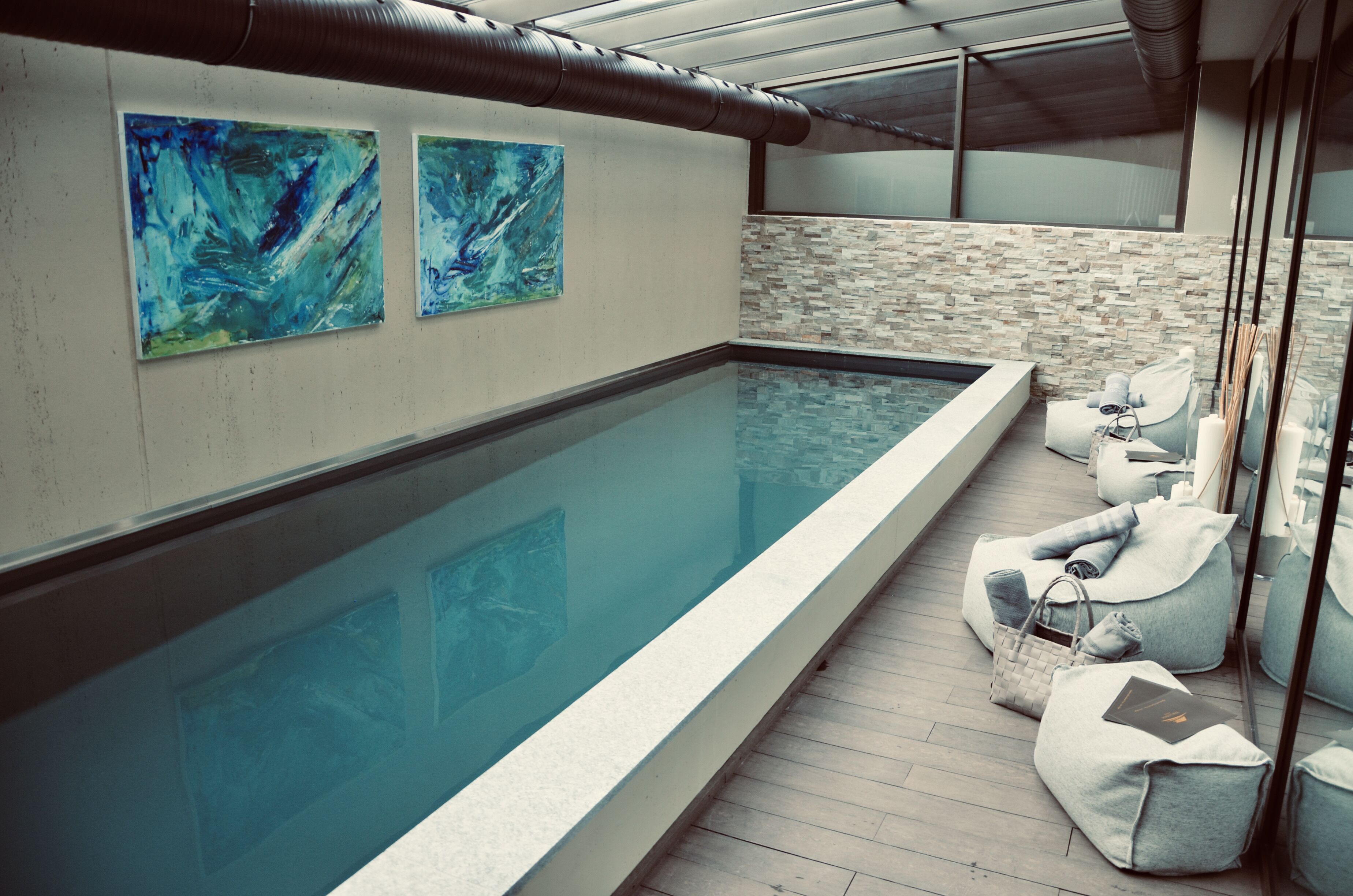 Una piscina interior moderna amb una paret de vidre, dues pintures abstractes i zones d'ambientació amb bosses.