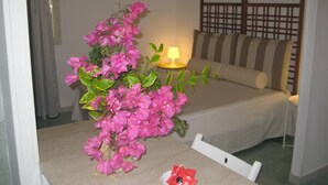 A hotel room with a bed, a bedside table with a lamp, a chair, and a vase with pink flowers.