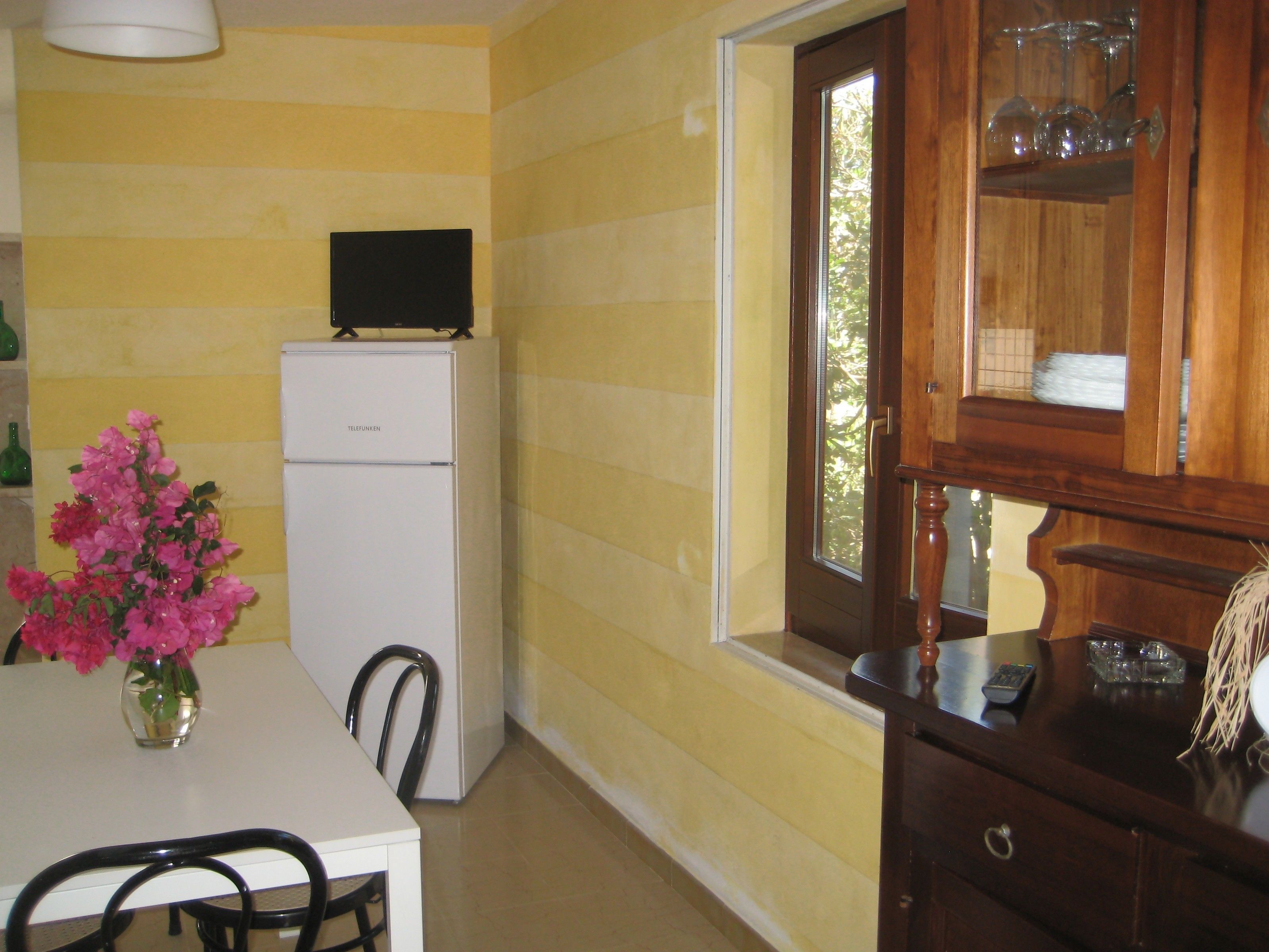 A kitchen with a white refrigerator, a dining table with chairs, a vase of pink flowers, and a wooden cabinet with glassware.
