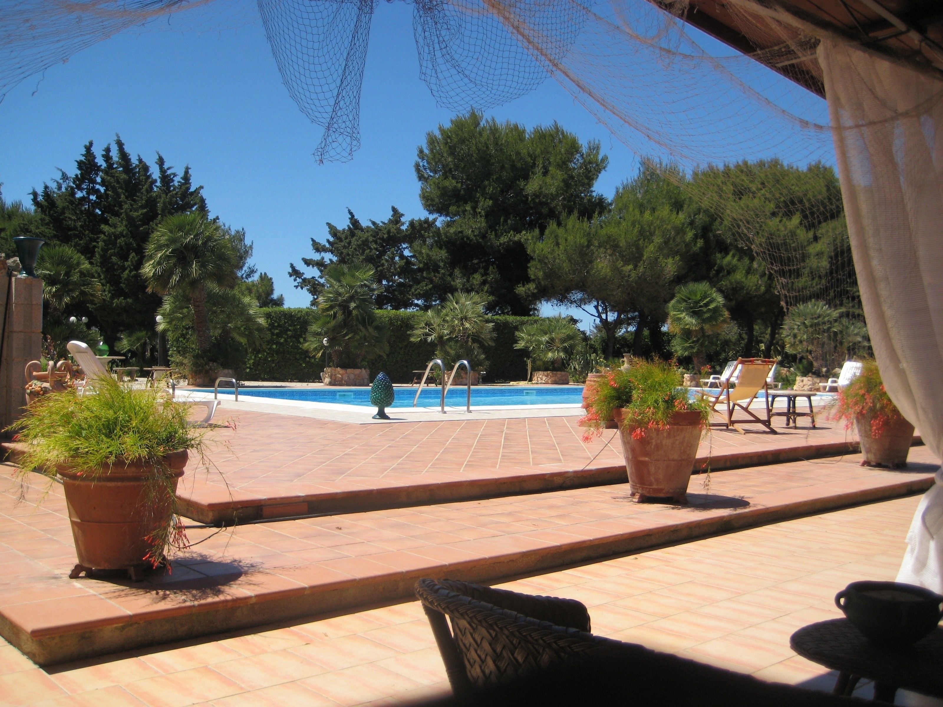 Un área de piscina con suelo de baldosas, sillones de descanso y plantas en macetas.