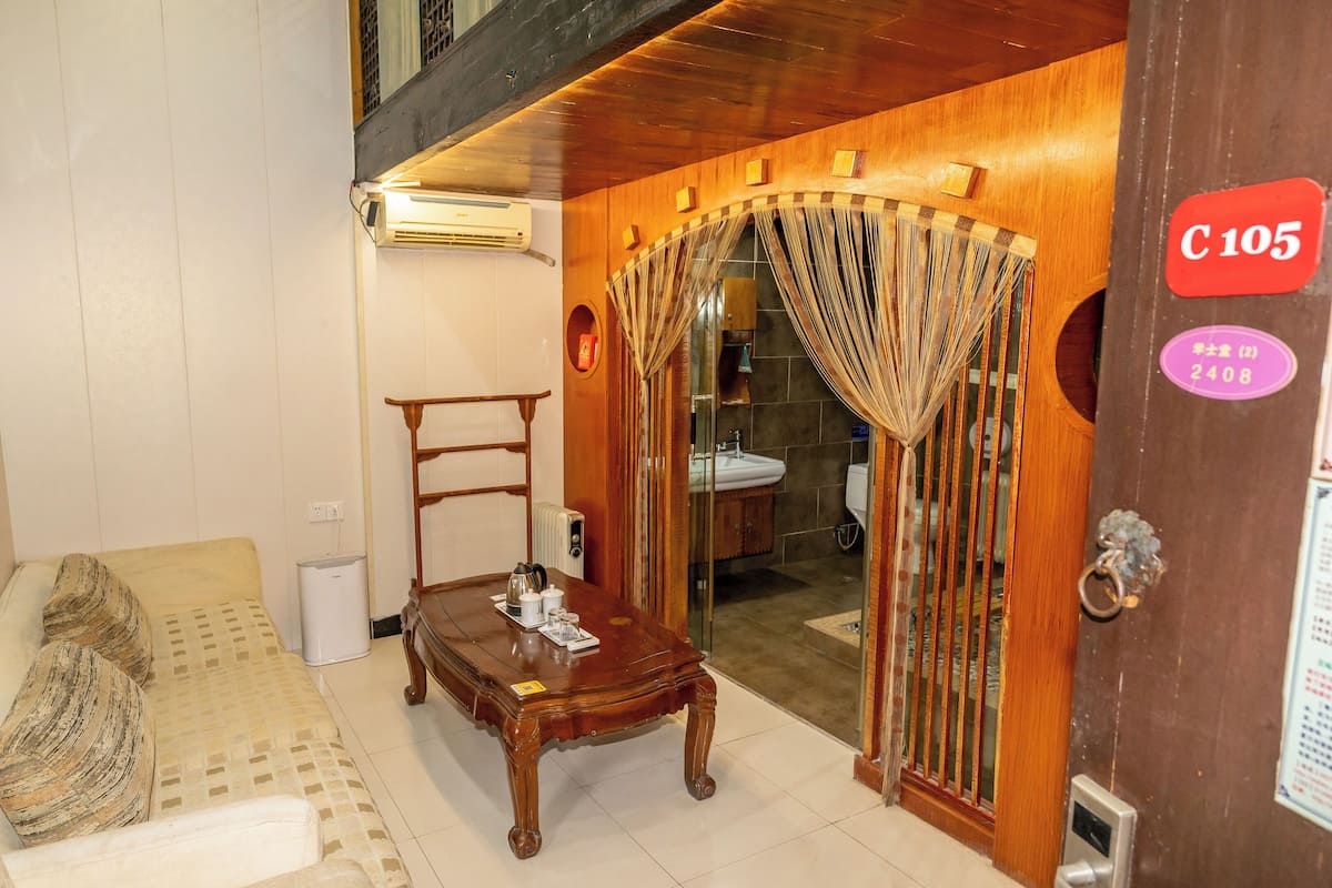 A hotel room with a bed, a wooden table with a tray of items, an air conditioning unit, and a bathroom visible through a glass door.