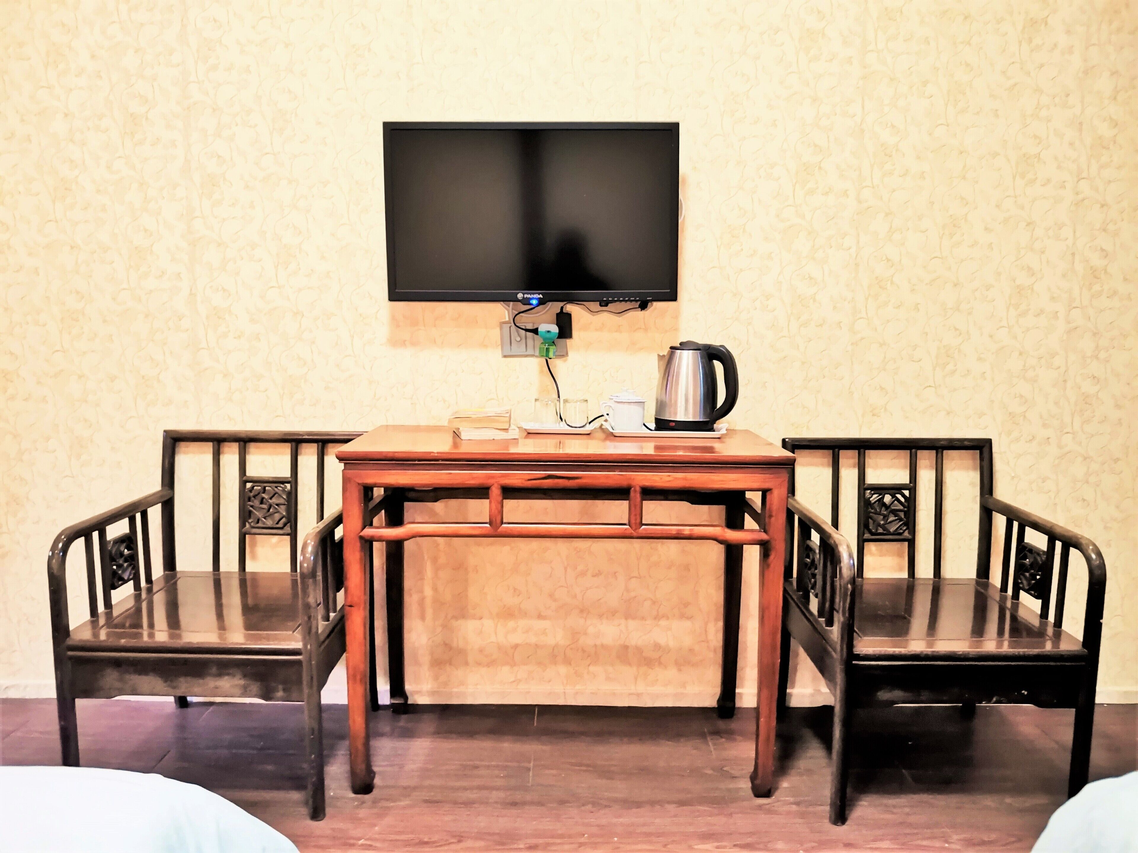 A dining area with a wooden table and chairs, a television mounted on the wall, and a kettle on the table.