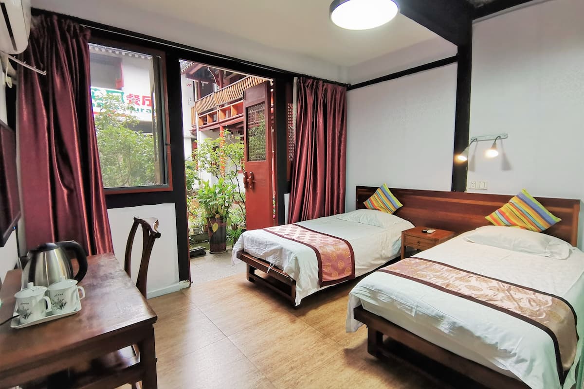 A hotel room with two beds, a wooden table with a kettle and teapots, a window with curtains, and a view of the outside balcony.