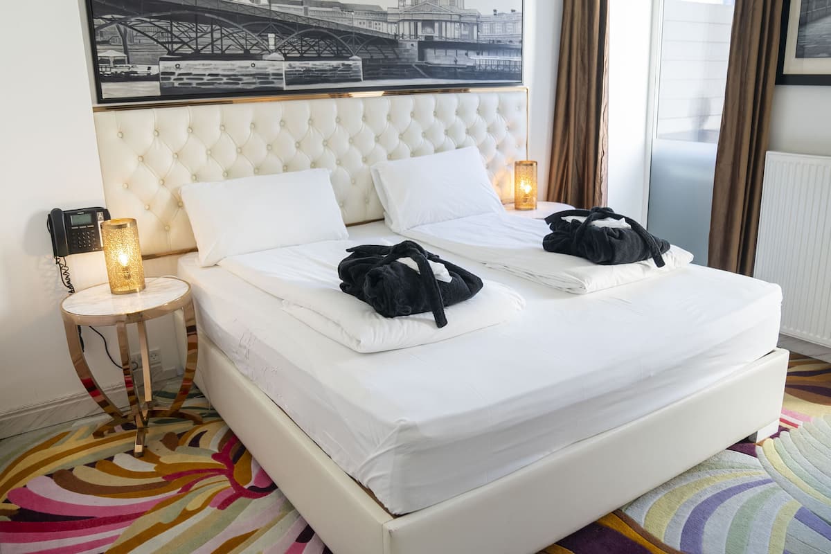 A hotel room with a white bed, two pillows, a bedside table with a lamp, a telephone, and a colorful rug.