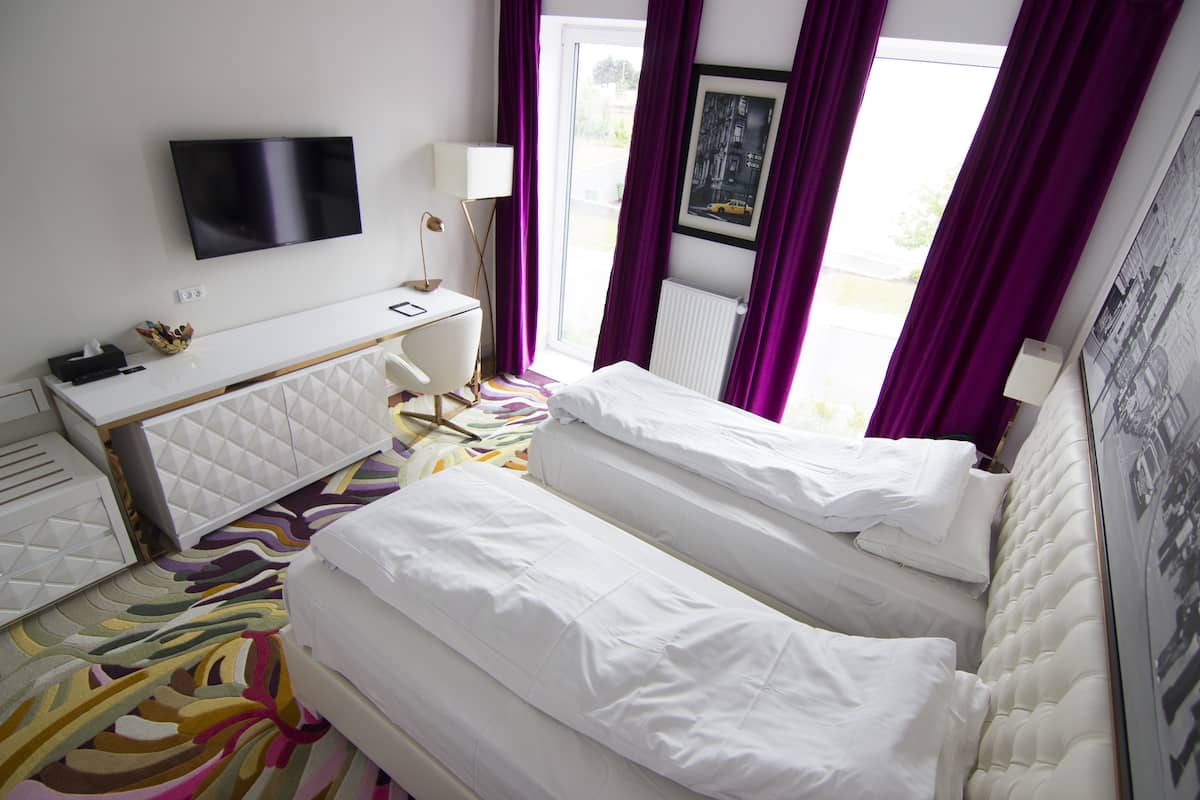 A hotel room with two beds, a desk, a TV, and a window with purple curtains.