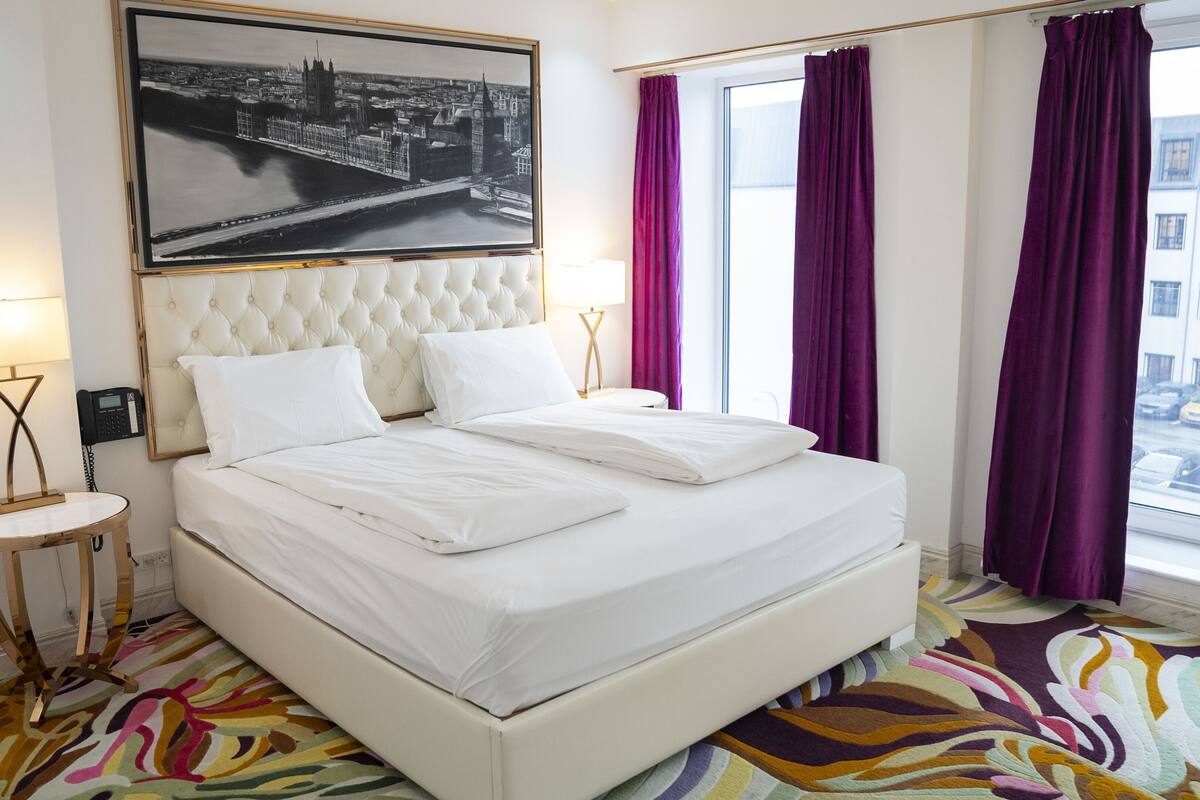 A hotel room with a large bed, a cityscape painting, a window with purple curtains, and a patterned carpet.