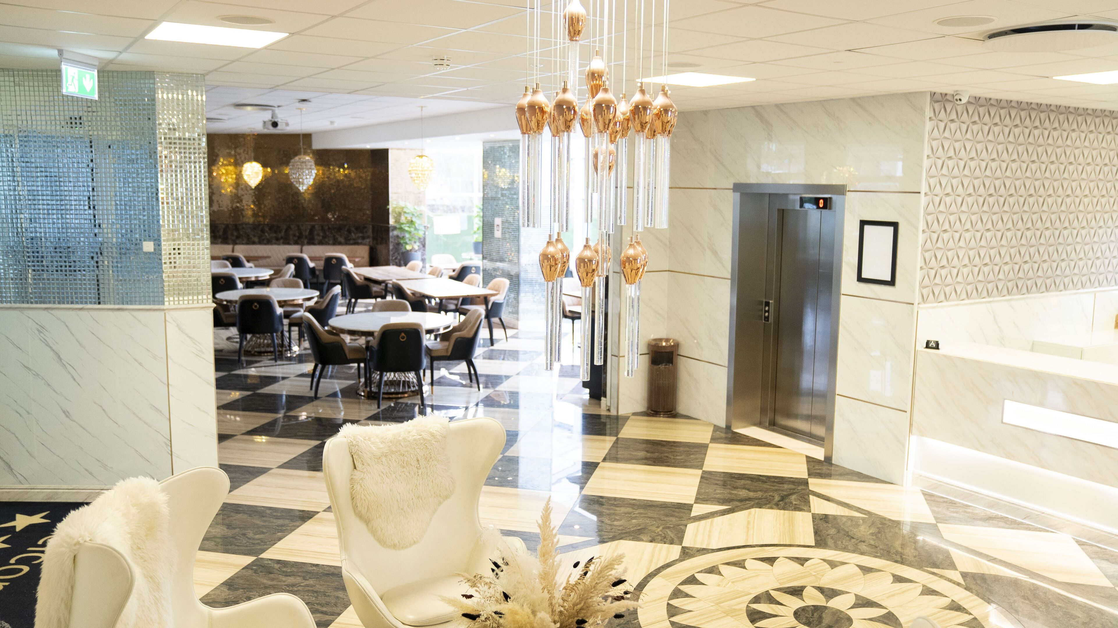 A hotel lobby with checkered flooring, a central seating area with chairs and tables, a decorative hanging light fixture, and a reception desk.