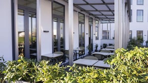 An outdoor seating area with tables and chairs, surrounded by potted plants and a building with large windows. - A Hotels Glostrup (Brondby)