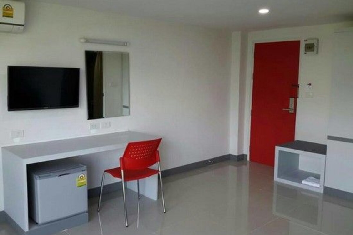 A hotel room with a white desk, a red chair, a television, a red door, and a small cabinet.