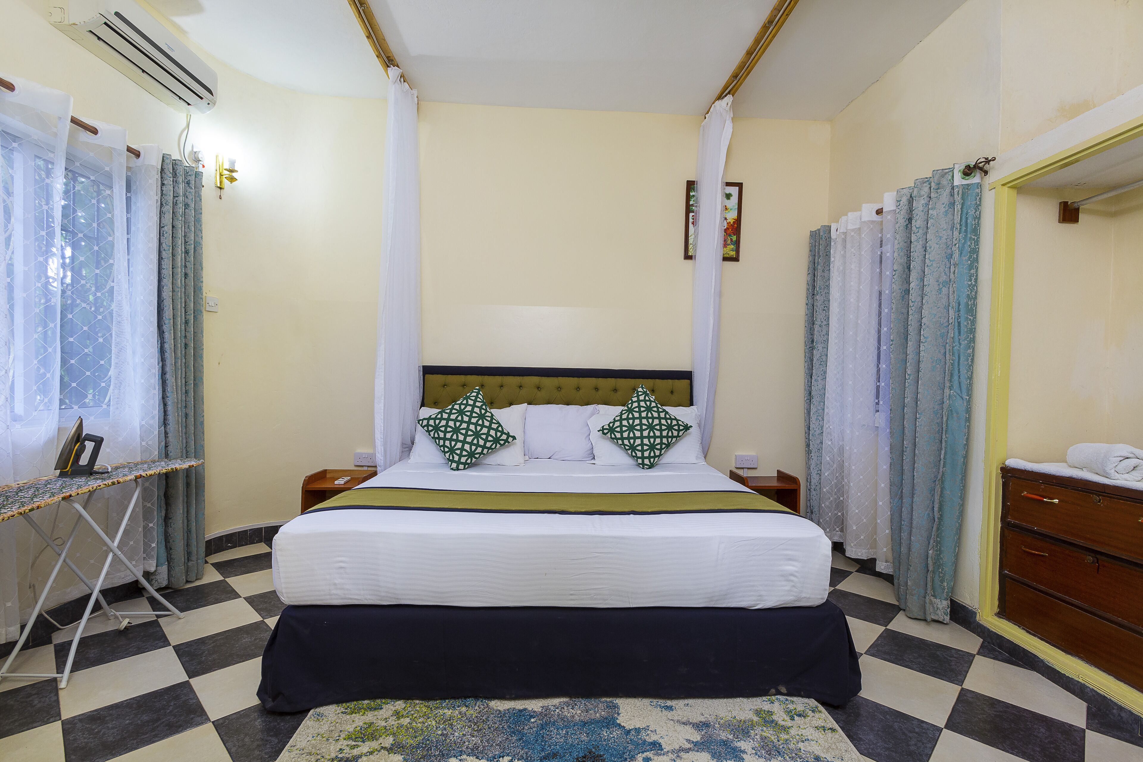 Una habitación de hotel con cama, mesita de noche, cómoda, tabla de planchar y cortinas.