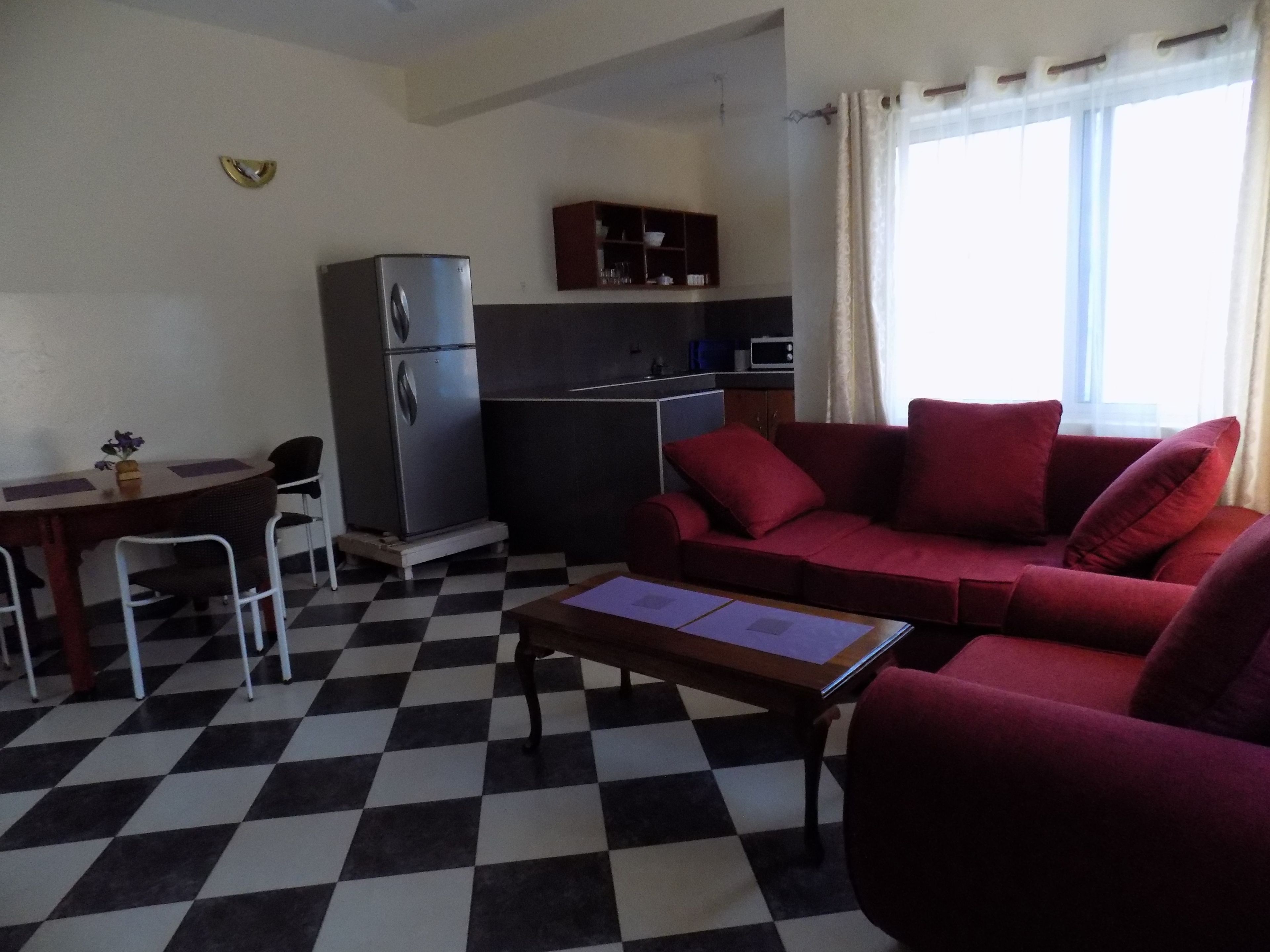 A compact living space with a checkered floor, a red sofa, a small dining table with chairs, a refrigerator, and a kitchenette area.