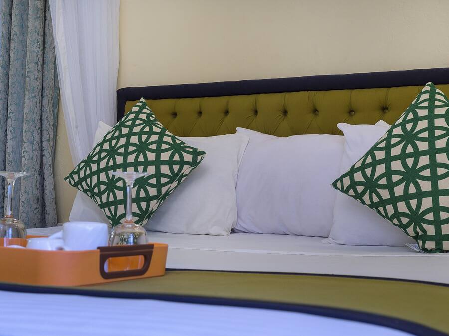 Quarto de hotel com cama, duas almofadas decorativas, um suporte com copos e uma xícara, e janela com cortinas.