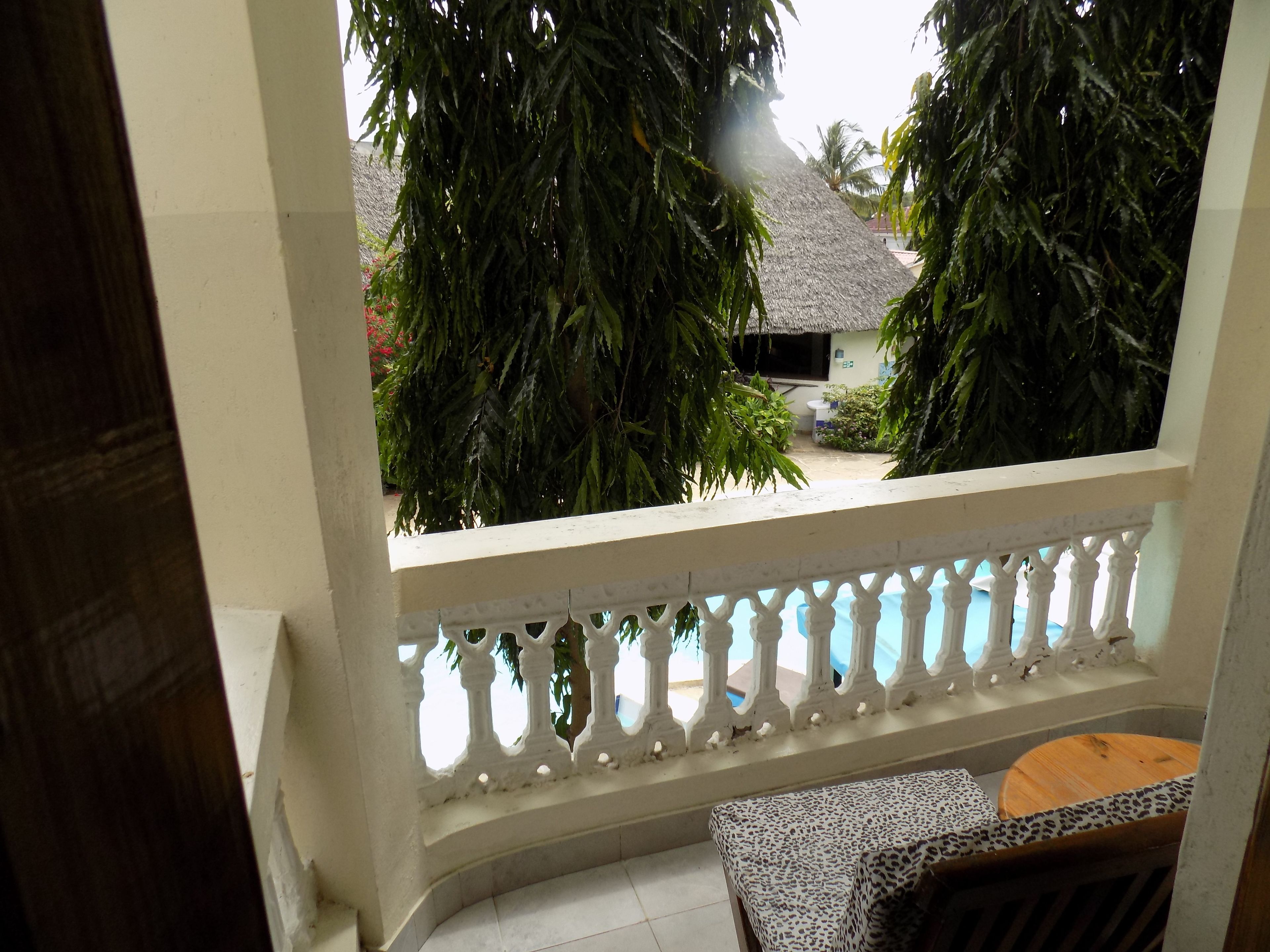 A balcony with a white balustrade, a small table with a patterned tablecloth, and a view of a swimming pool and greenery.