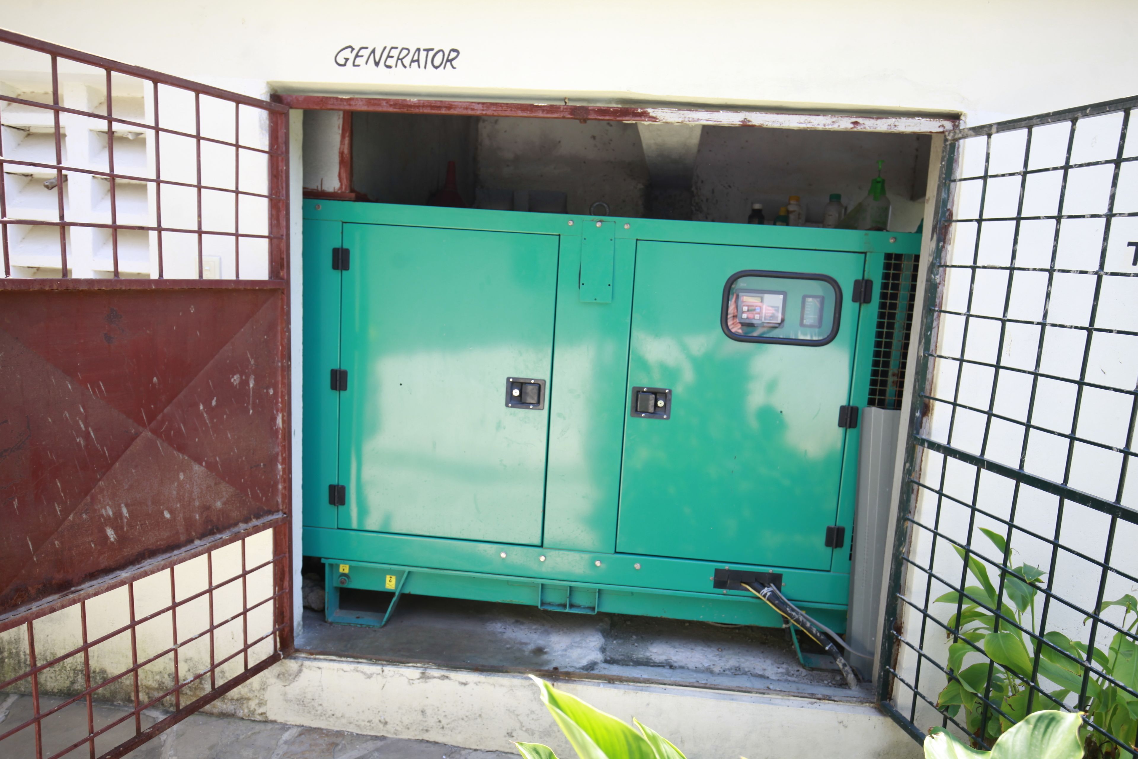 A green generator with a control panel, located in a corner with a metal gate and a concrete floor.