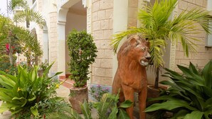 Un jardín con una estatua de león rodeado de diversas plantas y un edificio con columnas de piedra al fondo.