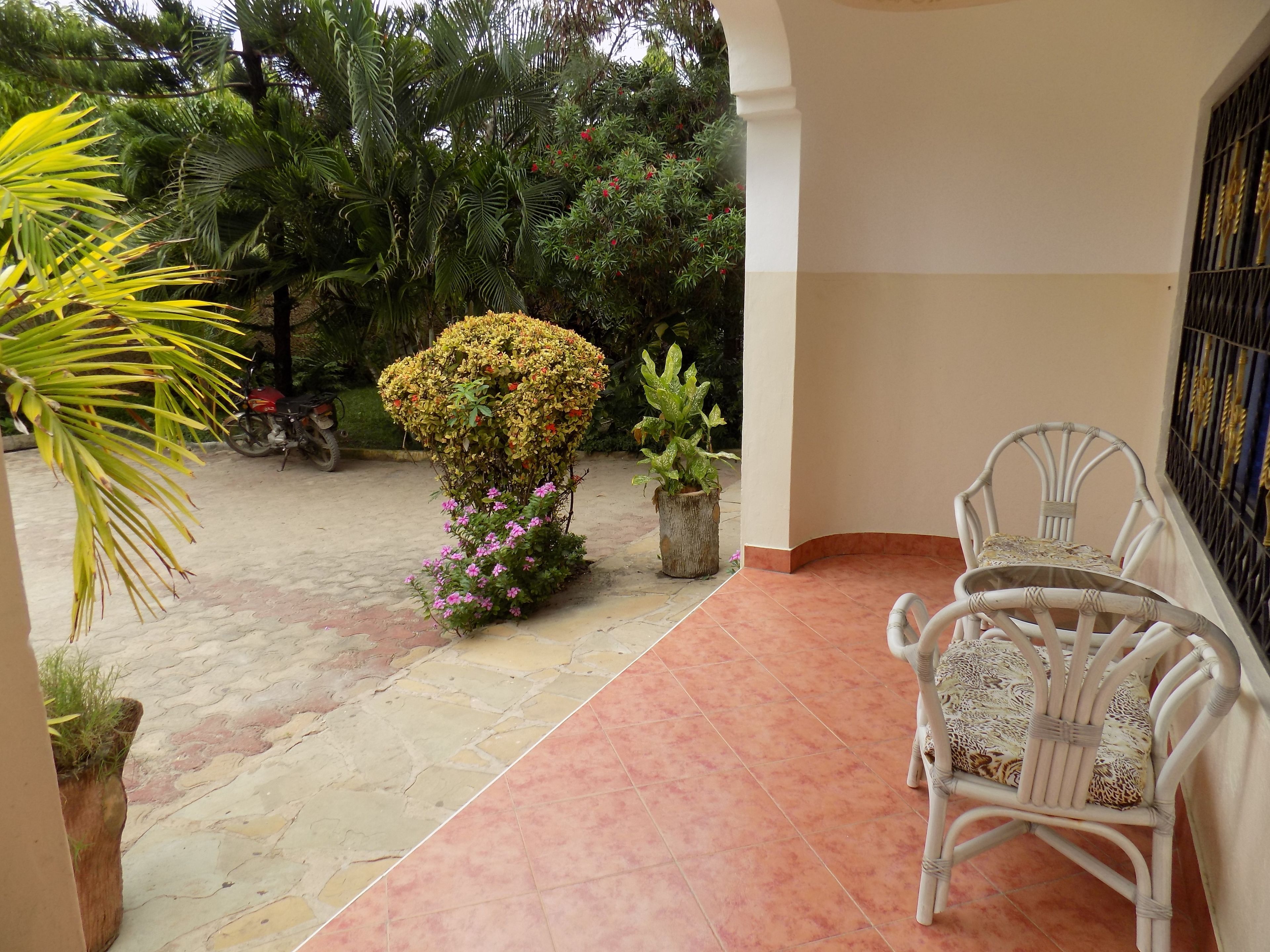 A covered patio area with tiled flooring, a white wicker chair, a potted plant, and a small tree.