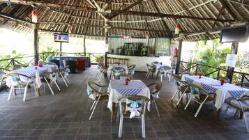 Een openluchtrestaurant met tafels en stoelen, een bar en een rieten dakconstructie.