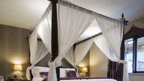 Uma cama com dossel e cortinas, roupa de cama branca com detalhes em roxo, duas mesas de cabeceira com luminárias e vista para o exterior através de uma janela.