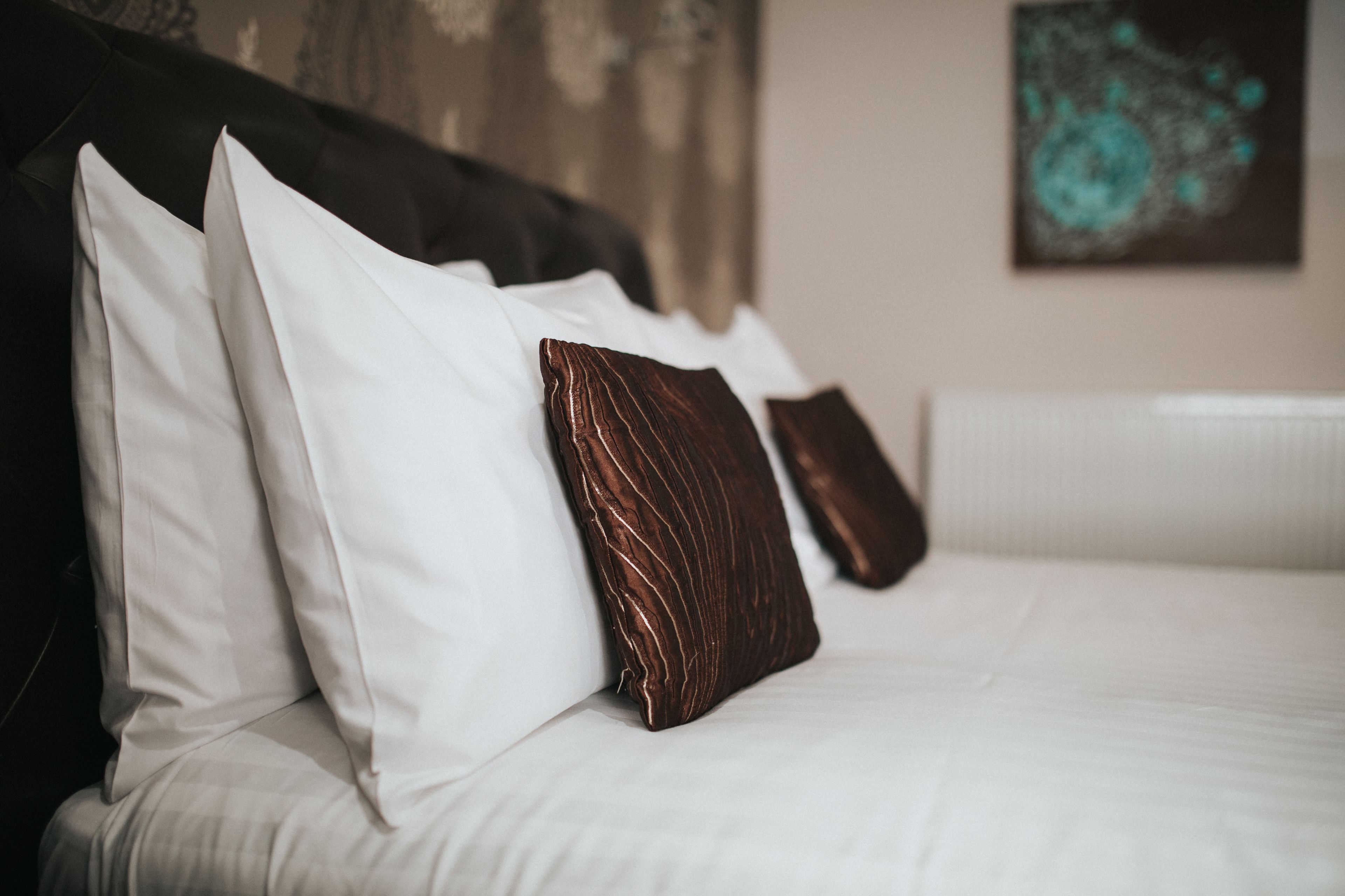 A neatly made bed with white and brown pillows, a dark headboard, and a teal abstract painting on the wall.