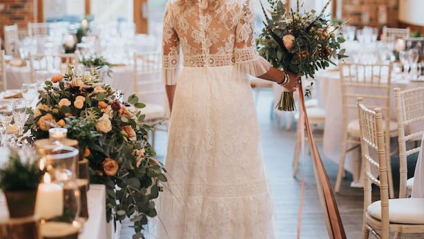 Uma noiva de vestido branco de renda segura um buquê, em um espaço com mesas preparadas para uma recepção de casamento.