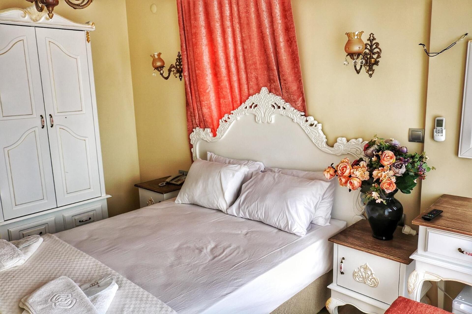 A neatly made bed with white linens, a vase of flowers on a bedside table, and a white wardrobe with a mirror.