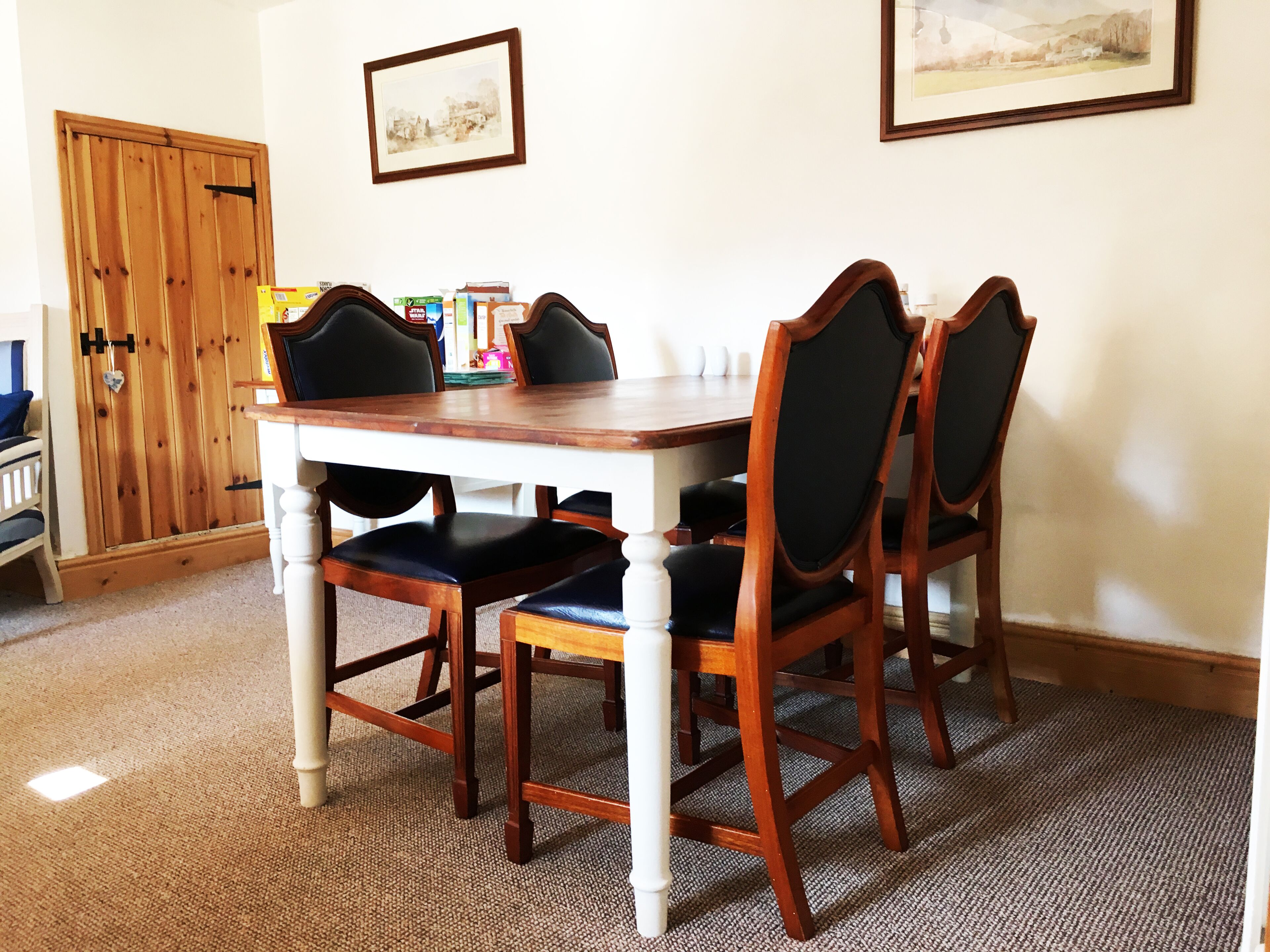 Un comedor con una mesa de madera y cuatro sillas, una puerta y dos cuadros en la pared.