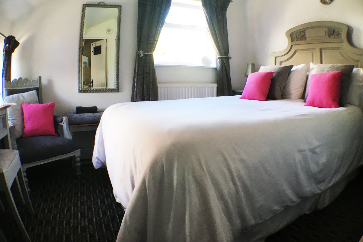 A bedroom with a large bed, a chair, a mirror, and a window with curtains.