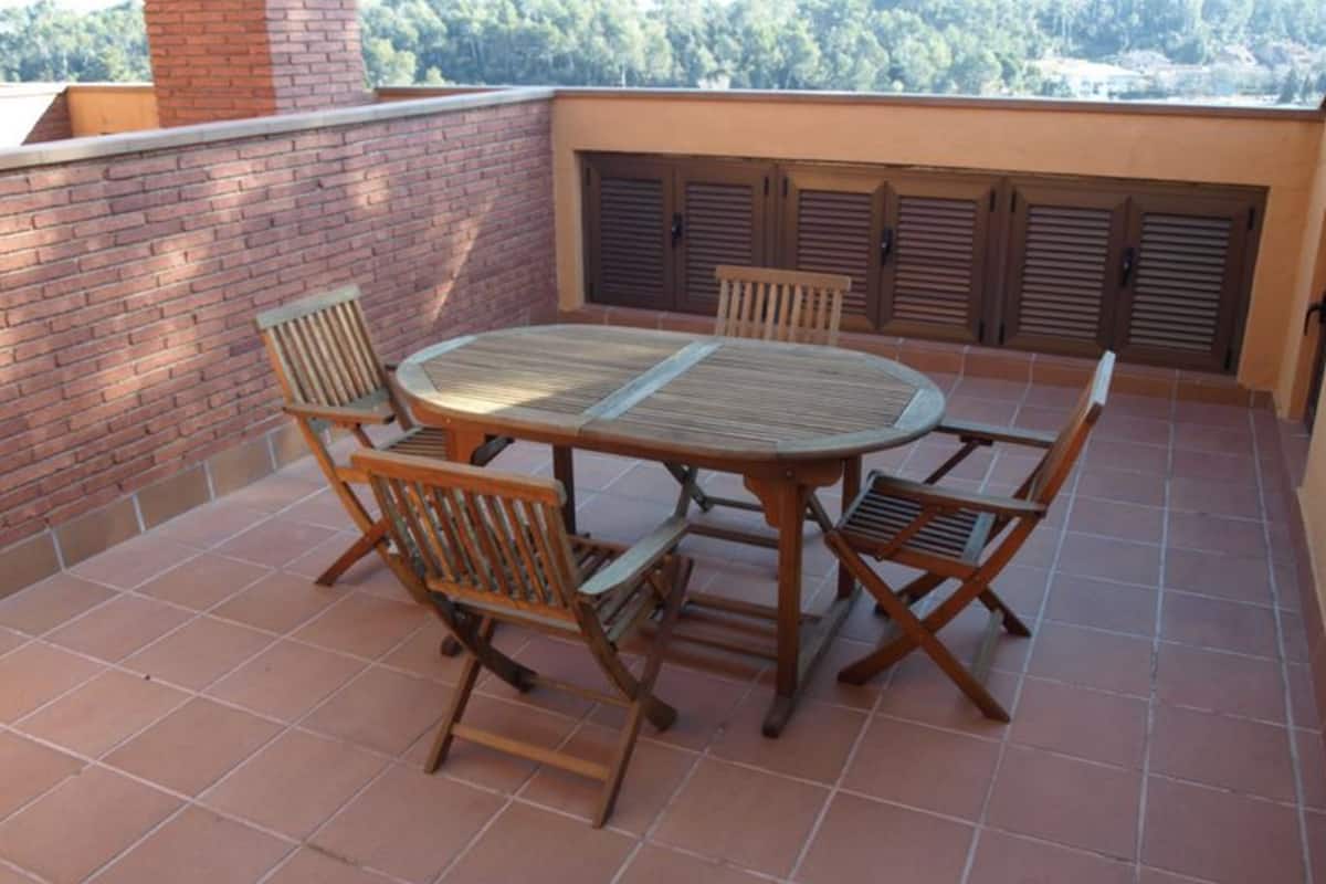 A patio with a round wooden table and six chairs, surrounded by a brick wall and a view of a forested area.