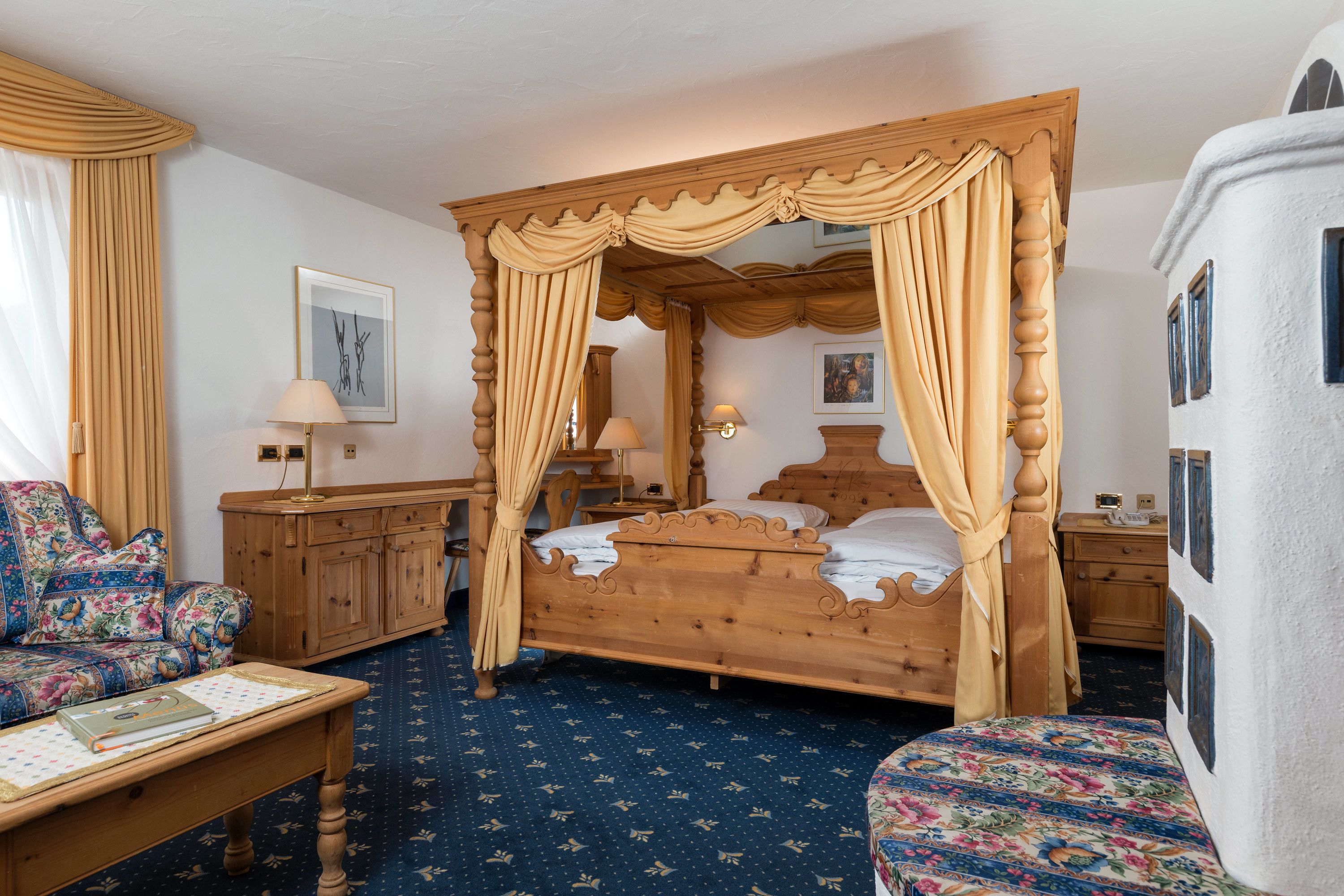 A traditional four-poster bed with curtains, a wooden nightstand, a floral sofa, and a matching armchair.