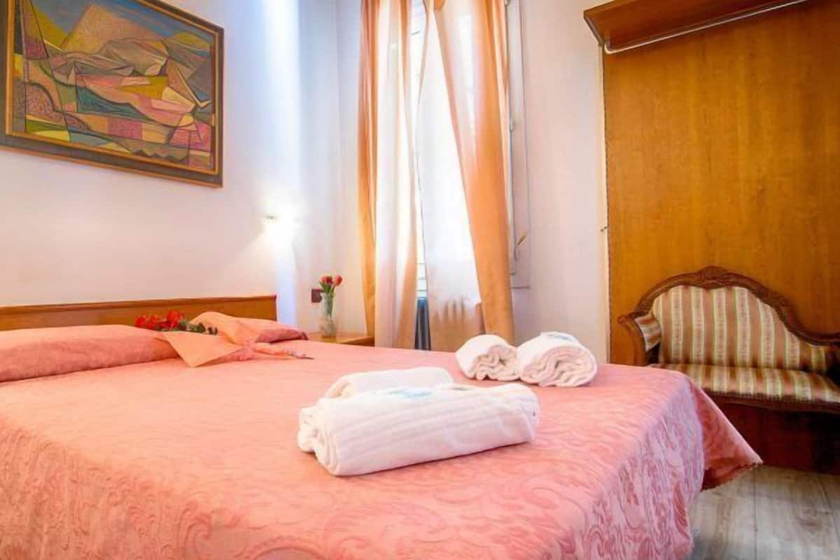 Un lit bien fait, avec une literie rose, un tableau accroché au mur et une chaise agrémentée d’un coussin à rayures.