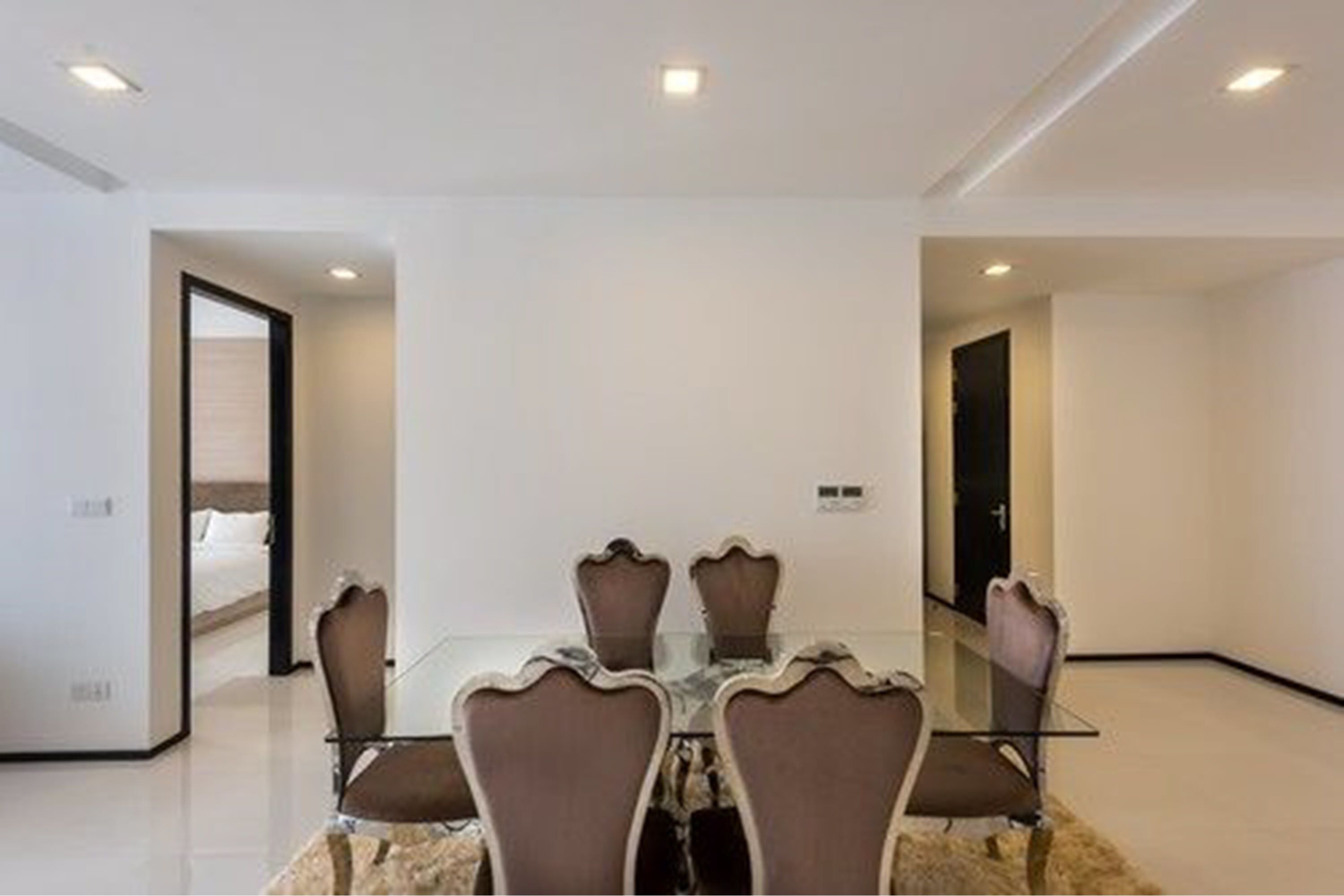 A modern hotel room with a glass dining table and four chairs, a large bed with white linens, and a door leading to another room.