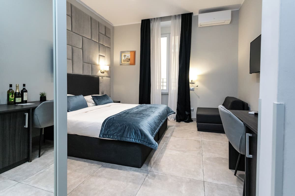 A modern hotel room with a bed, a desk, and a chair. There is a window with curtains, a picture on the wall, and a flat-screen TV mounted on the wall.