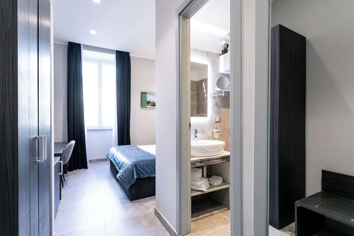 A modern bedroom with a bed, a desk, a chair, a bathroom with a sink and mirror, and a wardrobe.