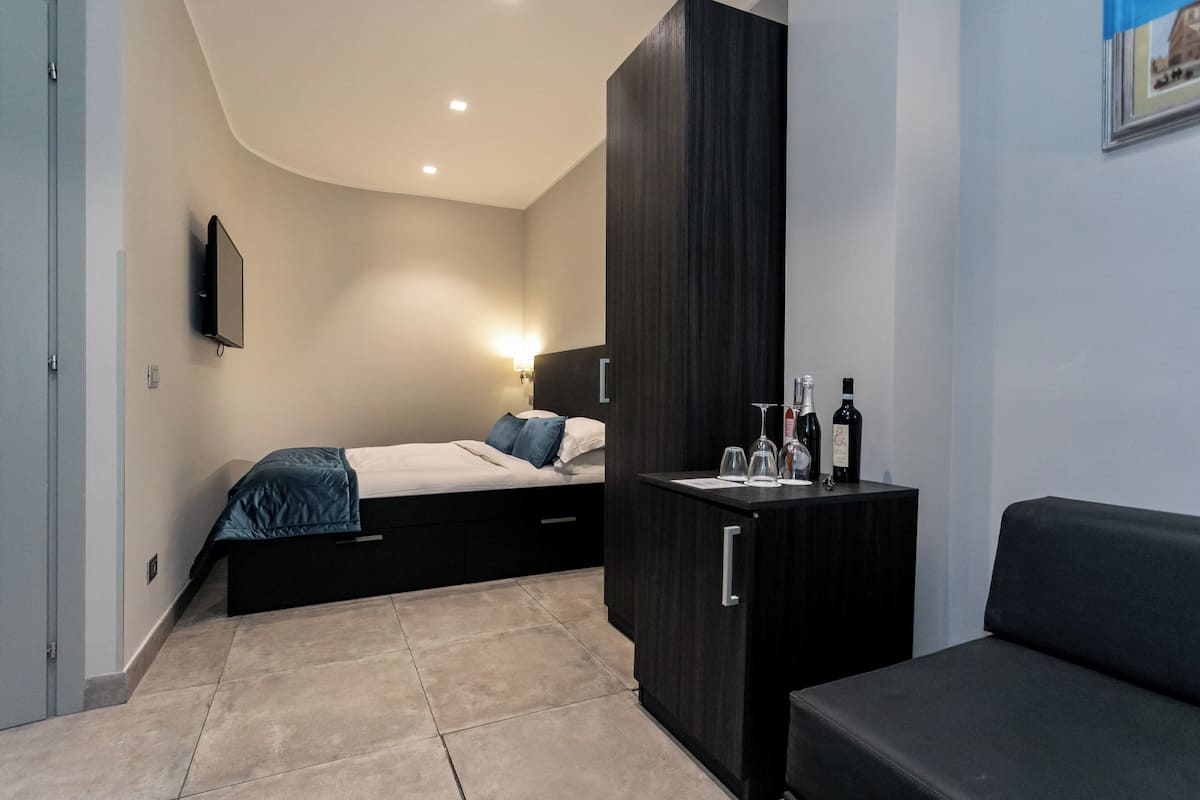 A modern bedroom with a bed, a wardrobe, a small table with bottles, and a TV mounted on the wall.