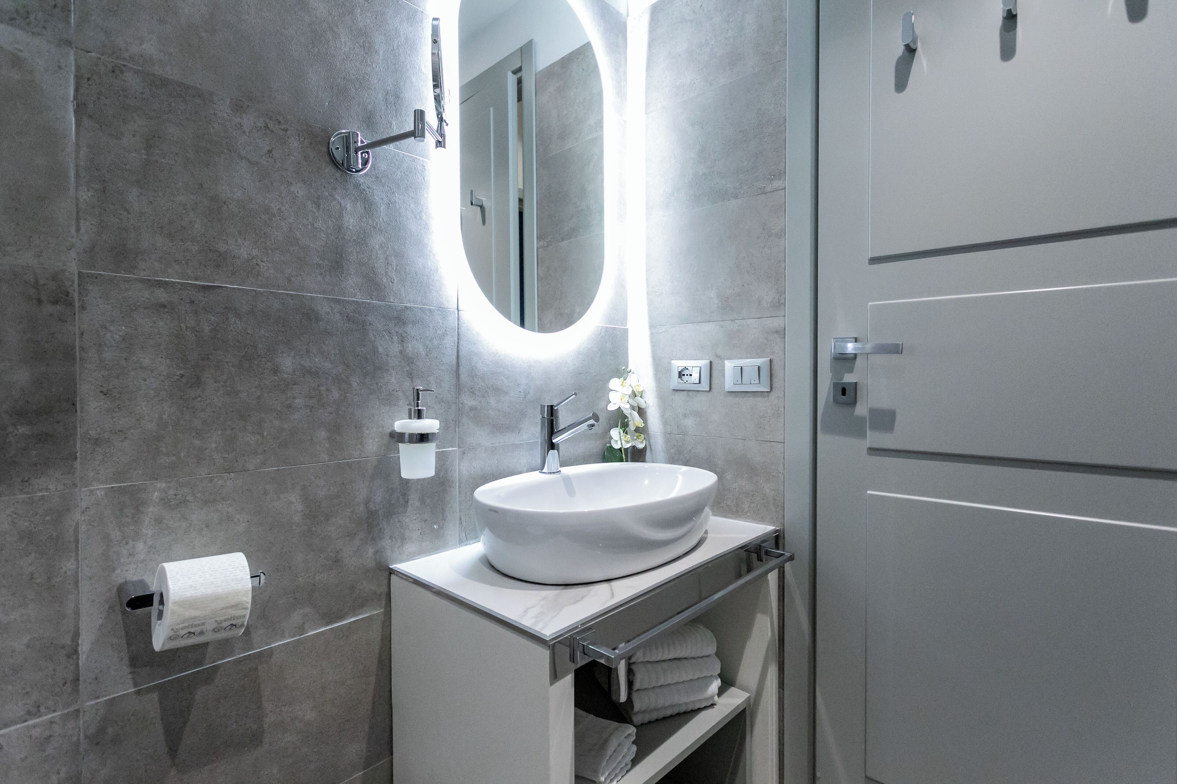 Un baño moderno con un lavamanos blanco, un espejo con luz circular y un perchero para toallas.