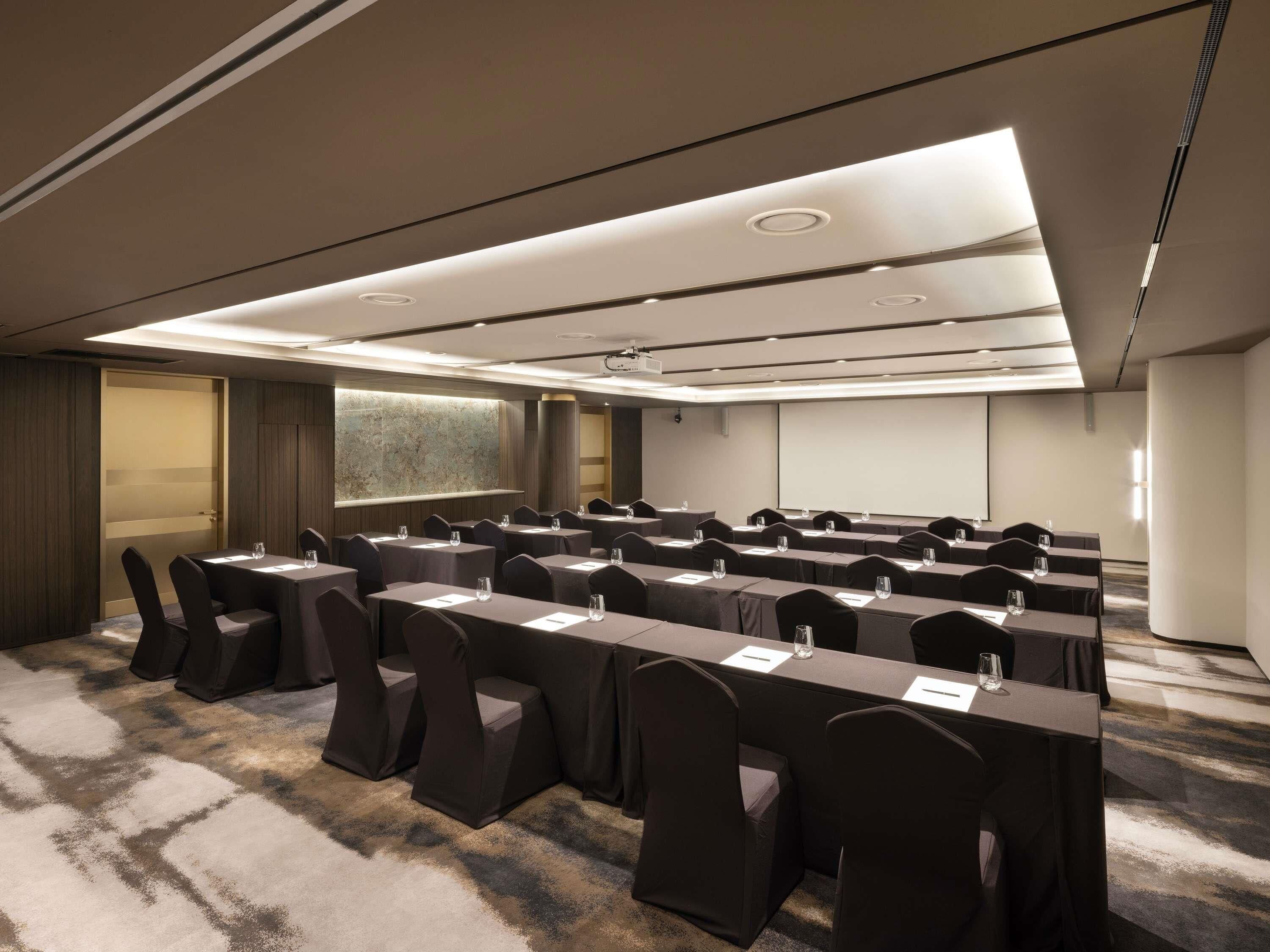 Un salón de conferencias con filas de mesas y sillas, una pantalla de proyector y piso alfombrado.