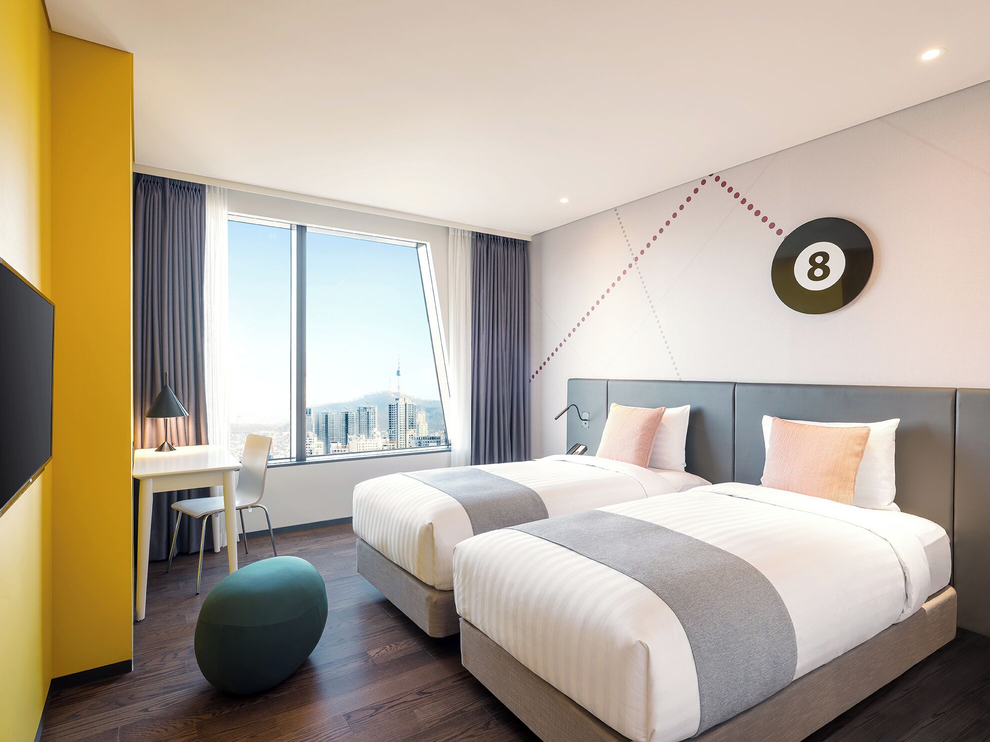 A hotel room with two beds, a desk, and a large window offering a city view.