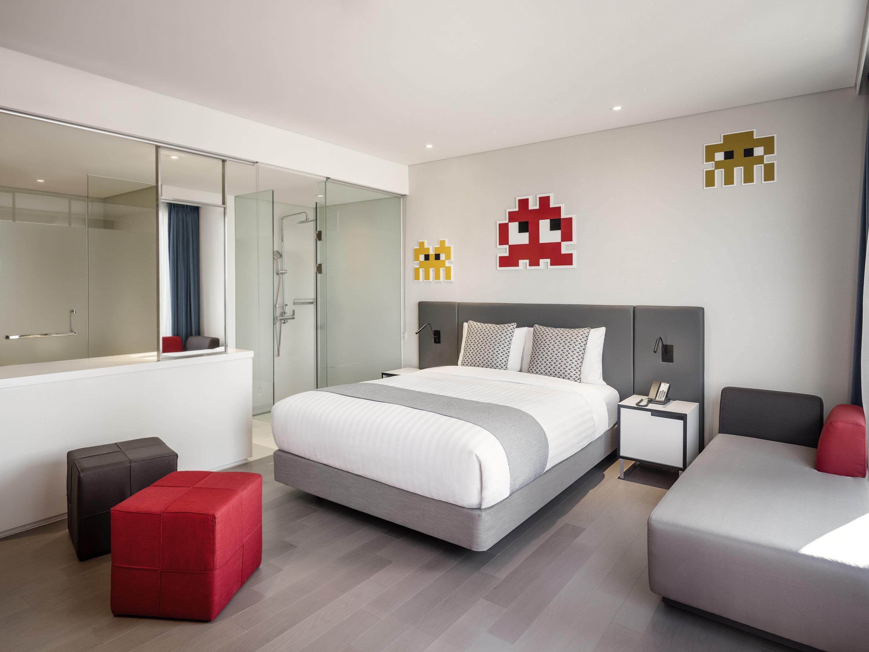 Una habitación de hotel moderna con una cama grande, cabina de ducha de vidrio, un pouf rojo y un sofá gris.