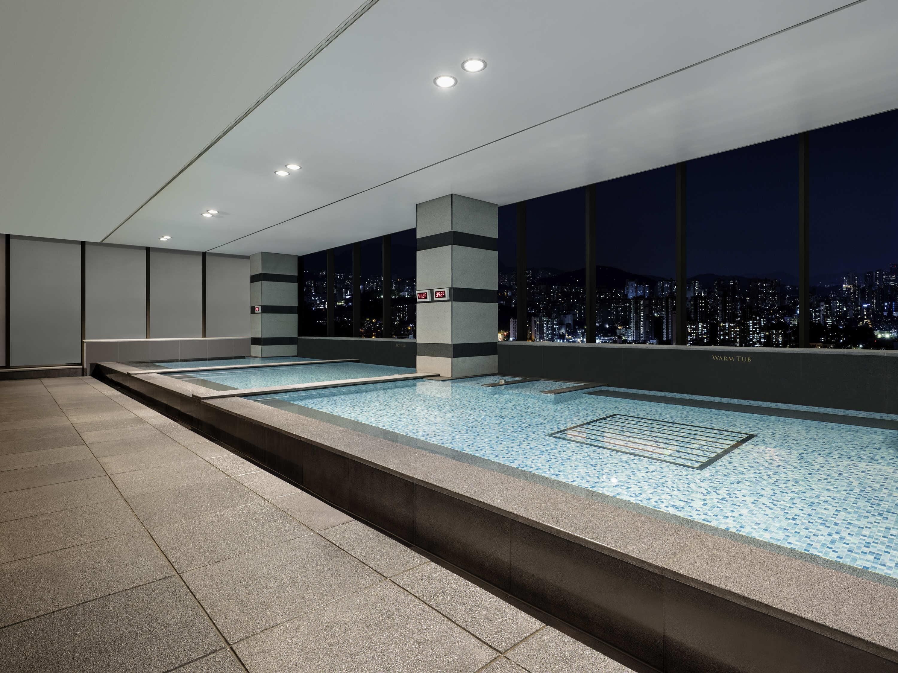 A modern indoor swimming pool with a city view, recessed lighting, and a tiled floor.