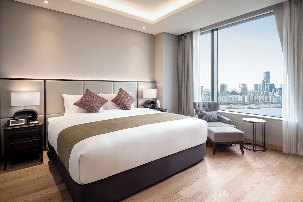 Une chambre d’hôtel moderne avec un grand lit, des tables de chevet, une chaise, une vue sur les toits de la ville par une grande fenêtre, et un panorama urbain visible à travers cette fenêtre.