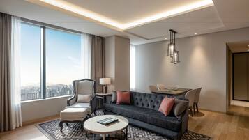 A modern living room with a sofa, armchair, and coffee table, large windows with a city view, and a dining area with a table and chairs.